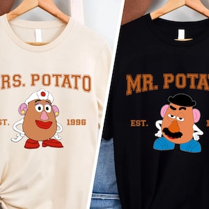 May include: Two t-shirts, one beige and one black, with a cartoon image of Mr. and Mrs. Potato Head. The beige shirt says "MRS. POTATO" with "EST. 1996" below the image. The black shirt says "MR. POTATO" with "EST. 1995" below the image.