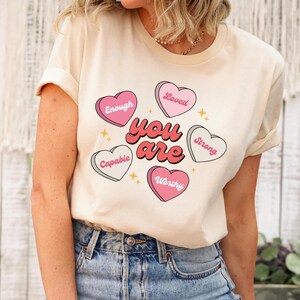 May include: Cream-colored t-shirt with the words "You Are" in red and white, surrounded by pink and white candy hearts. Each heart has a positive word: Enough, Loved, Capable, Strong, and Worthy. Gold star accents.