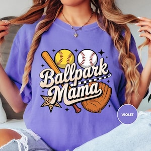 A woman wearing a purple t-shirt with a graphic design of baseball equipment and the text "Ballpark Mama" is sitting on a bed.
