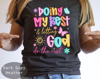 May include: Dark grey heather t-shirt with the text "Doing My Best & Letting God Do The Rest" in colorful, playful lettering. The design includes butterflies, flowers, and a sun graphic. A casual, inspirational tee.