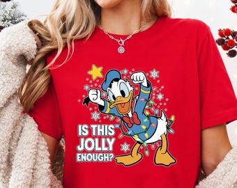 May include: A red t-shirt featuring a cartoon Donald Duck character decorated with Christmas lights and snowflakes. The text "IS THIS JOLLY ENOUGH?" is printed below the character. The shirt is a festive holiday design.
