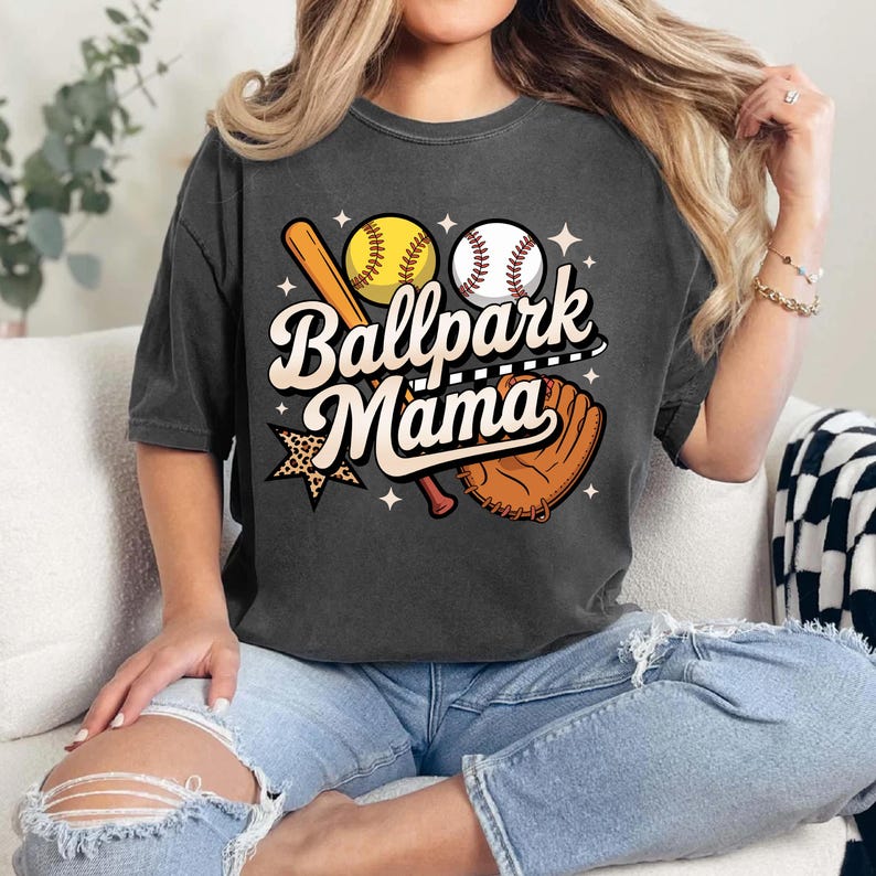 A woman wearing a gray t-shirt with a graphic design of baseball equipment and the text "Ballpark Mama" is sitting on a couch.