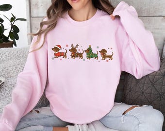 May include: A light pink sweatshirt featuring a festive dachshund design. The graphic includes four dachshunds dressed in holiday attire: a Santa suit, reindeer antlers, a Christmas tree, and a gingerbread outfit. Snowflakes are scattered around the dogs.