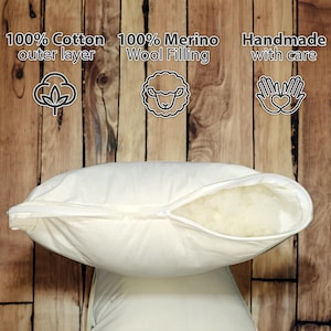May include: A white pillow with a zipper opening, showing the white merino wool filling. The pillow is made with a 100% cotton outer layer and is handmade with care. The pillow is on a wooden surface.