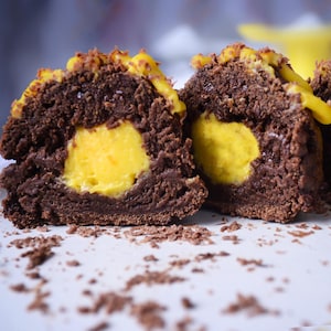 Könnte beinhalten: Drei Schokoladen-Cake-Pops mit einer gelben Füllung, halbiert, die die Füllung freilegt. Die Cake-Pops sind mit Schokolade überzogen und haben einen gelben Überzug. Die Cake-Pops liegen auf einem weißen Teller mit Schokoladenspänen.