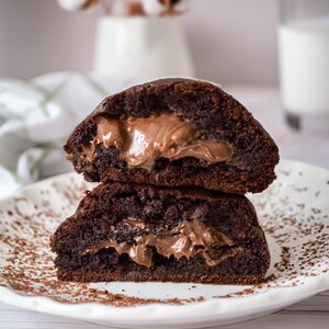 Puede incluir: Dos mitades de una galleta de chocolate con un relleno cremoso marrón claro, apiladas en un plato blanco con motas marrones. Las galletas tienen un exterior brillante de color marrón oscuro. Un vaso de leche y un jarrón con flores de algodón están en el fondo.