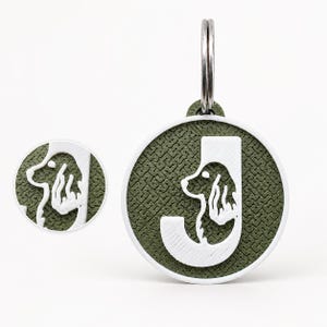 Pode incluir: Uma placa circular para animais de estimação com a letra J branca integrada numa silhueta branca de um cão spaniel. O fundo tem um padrão texturado verde-azeitona, emoldurado a branco com um anel de metal prateado no topo.