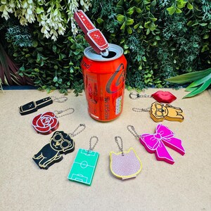 Op de afbeelding: Een verzameling sleutelhangers in diverse vormen, waaronder een rode glitterlippenstift, een rode roos, een zwarte hond met een bril, een groen voetbalveld, een roze kattenkop, een grote roze strik, een gele hond en rode lippen.