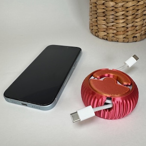 May include: A red cable organizer with a white cable wrapped around it. The organizer has a ribbed texture and a circular shape. A black smartphone is in the background.