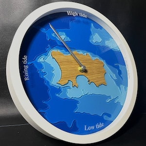 Op de afbeelding: Een ronde getijdenklok met een witte lijst, met een blauw en houten kaartontwerp. De wijzerplaat toont de diepten van de oceaan en een houten eiland. De wijzers geven "Hoogwater" en "Laagwater" aan.