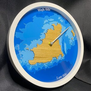 Può includere: Un orologio di marea circolare con cornice bianca che presenta una massa terrestre centrale color marrone chiaro circondata da profondità d'acqua blu a strati. Il quadrante include il testo High tide, Rising tide, Low tide e Falling tide con una lancetta argentata.