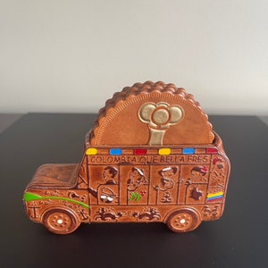 May include: Brown ceramic coaster holder shaped like a bus, with a stack of round coasters. The bus has painted details, including the text "COLOMBIA QUE BELLA FRES" and images of people. The coasters are brown with a gold design.
