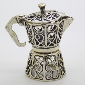 May include: A silver-colored, filigree-style coffee maker. The decorative espresso pot features an ornate, openwork design on the body, lid, handle, and spout. The handle is curved, and the lid has a small knob.