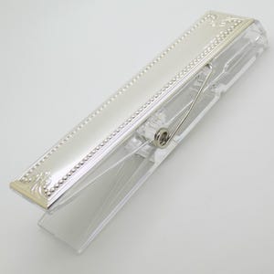 May include: A clear plastic clothespin with a silver-colored, rectangular top. The top has a decorative border with small, raised dots and ornate corner designs. A silver-colored spring and wire mechanism are visible.