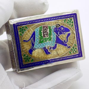 Puede incluir: Una caja rectangular plateada con un diseño ornamentado. La tapa presenta un elefante azul con una manta estampada, sobre un fondo dorado. El borde es azul con detalles florales. La caja está sujeta por una mano enguantada.