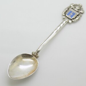 May include: Silver spoon with a decorative handle and a blue and white enamel crest. The spoon has a rounded bowl and a long, slender handle. The crest features a shield design. A collectible souvenir or decorative item.
