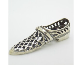 May include: A silver-colored shoe charm with an openwork design. The charm features a lattice pattern on the vamp and sides, with a small heel and a strap across the instep. The shoe charm is likely a pendant or a decorative element.