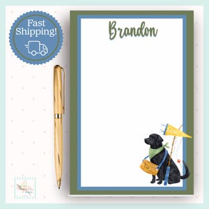 May include: A personalized notepad with a green and blue border, featuring the name "Brandon" in script. A watercolor illustration of a black Labrador with fishing gear is at the bottom. A gold pen rests on the side. A blue circle with "Fast Shipping!" is in the corner.