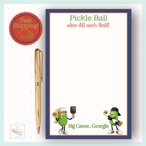 Op de afbeelding: Een notitieblok met een blauwe en rode rand met de tekst "Pickle Ball where dill meets thrill!" en cartoon augurken personages. Een gouden pen ligt naast het notitieblok. Het notitieblok bevat ook de tekst "Big Canoe, Georgia".