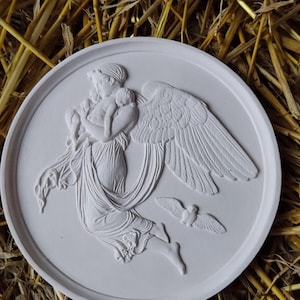 May include: A white, circular relief sculpture depicting a classical angel figure holding a child, with a dove in flight. The angel has large wings and flowing drapery. The sculpture is set against a background of straw.