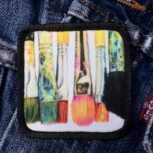 May include: A close-up of a patch with a design of five paintbrushes with colorful paint on them. The paintbrushes are arranged vertically and are all different sizes. The patch is white with a black border.