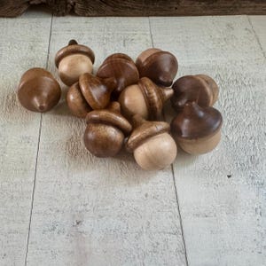 May include: A collection of wooden acorns in various shades of brown and tan. The acorns are handcrafted, with detailed caps and smooth, rounded bodies. The natural wood grain is visible, adding to their rustic charm.