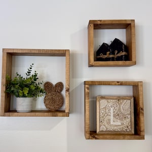 May include: Three square wooden shelves mounted on a white wall. One shelf holds a potted plant and a woven bunny. Another shelf displays two black house-shaped decorations. The third shelf contains a framed artwork with a paisley design.