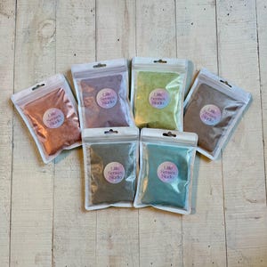 May include: Six small, clear plastic pouches filled with colored powders are arranged on a light wood-grain surface. Each pouch has a circular sticker with the text "Little Senses Studio". The powders are in shades of orange, purple, green, brown, gray, and teal.