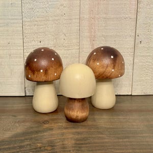 May include: Three wooden mushroom figurines. Two mushrooms have brown caps with white polka dots and cream-colored stems. The third mushroom has a cream-colored cap and a brown stem. The mushrooms are arranged on a wooden surface.