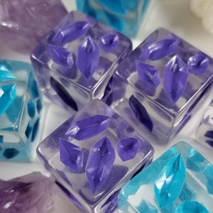 May include: Close-up of several clear resin dice with embedded purple and turquoise crystal shapes. The dice are square and have a glossy finish. Some amethyst crystals are also visible.
