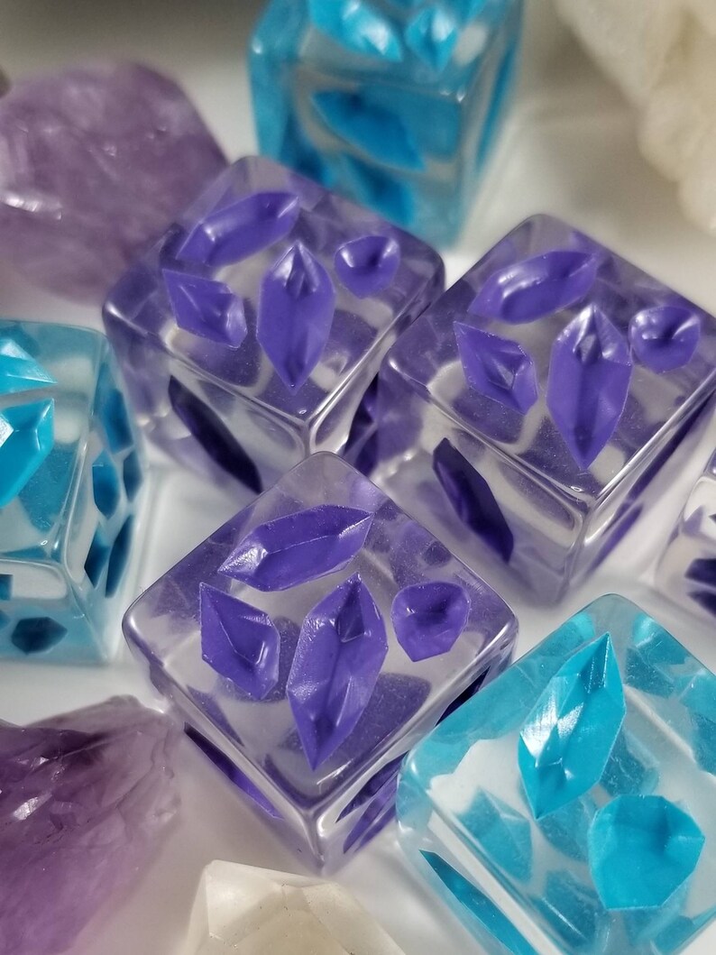 May include: Close-up of several clear resin dice with embedded purple and turquoise crystal shapes. The dice are square and have a glossy finish. Some amethyst crystals are also visible.