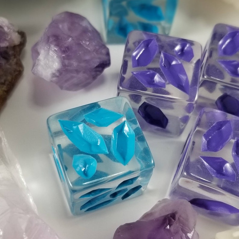 May include: Close-up of several clear dice with embedded blue and purple crystal shapes. The dice are arranged with raw amethyst crystals. The dice have a translucent appearance, and the crystals are a vibrant blue and purple.