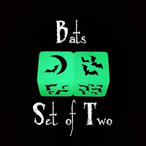 May include: Two green glow-in-the-dark dice with black bat silhouettes. One die has a crescent moon and bats, the other has multiple bats. The dice are labeled "Bats Set of Two".