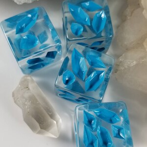 May include: Four clear, square dice with bright blue crystal-shaped designs inside. A clear quartz crystal is next to the dice. The dice are on a white surface, with a background of white and clear crystals.