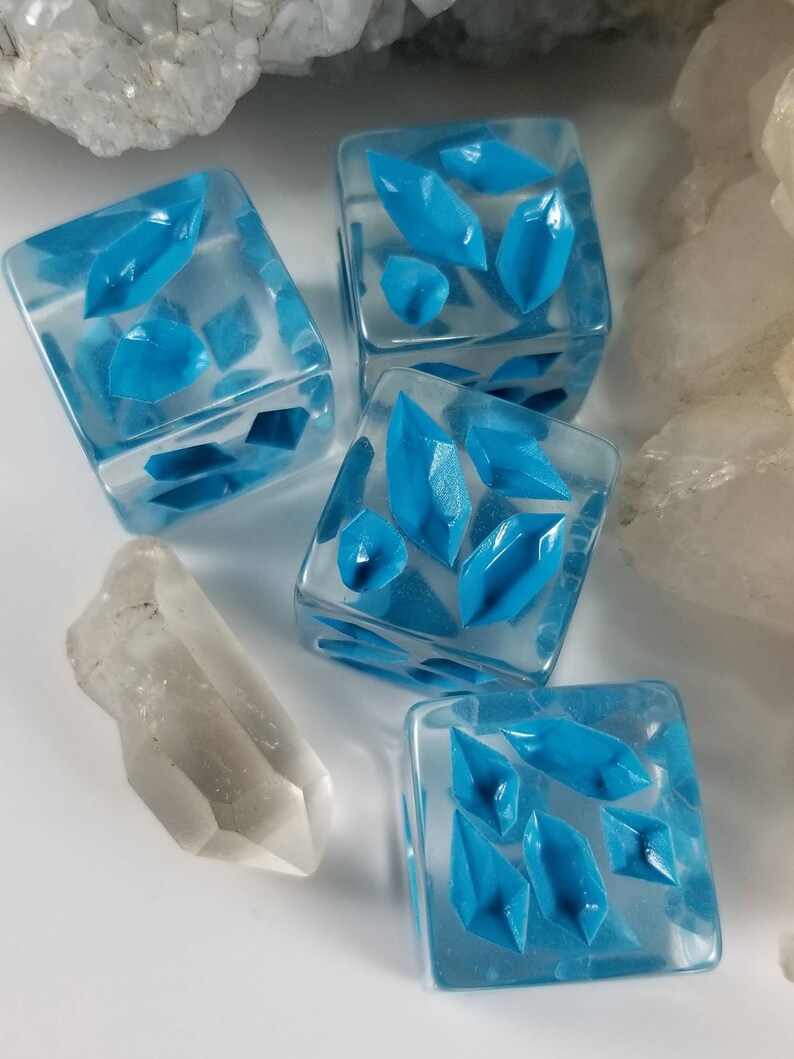 May include: Four clear, square dice with bright blue crystal-shaped designs inside. A clear quartz crystal is next to the dice. The dice are on a white surface, with a background of white and clear crystals.