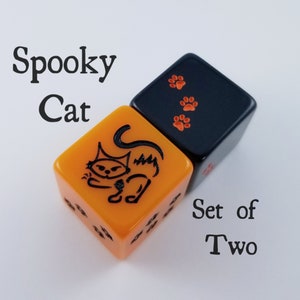 Peut inclure: Ensemble de deux dés, l'un orange avec une illustration de chat noir et l'autre noir avec trois empreintes de pattes orange. Les dés sont étiquetés "Spooky Cat" et "Set of Two".