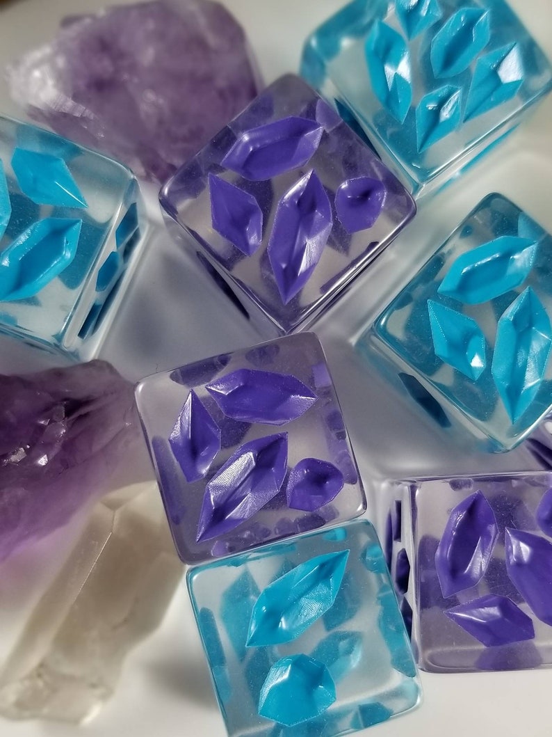 May include: Close-up of several clear, square resin dice with embedded purple and teal crystal shapes. Amethyst and clear quartz crystals are in the background. The dice are a unique, handmade item.