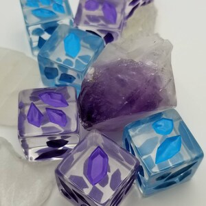 May include: A collection of clear, square dice with embedded purple and blue geometric shapes. The dice are arranged with a large, rough amethyst crystal and white, irregular stones. The dice have a translucent appearance.