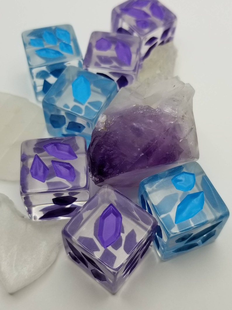 May include: A collection of clear, square dice with embedded purple and blue geometric shapes. The dice are arranged with a large, rough amethyst crystal and white, irregular stones. The dice have a translucent appearance.
