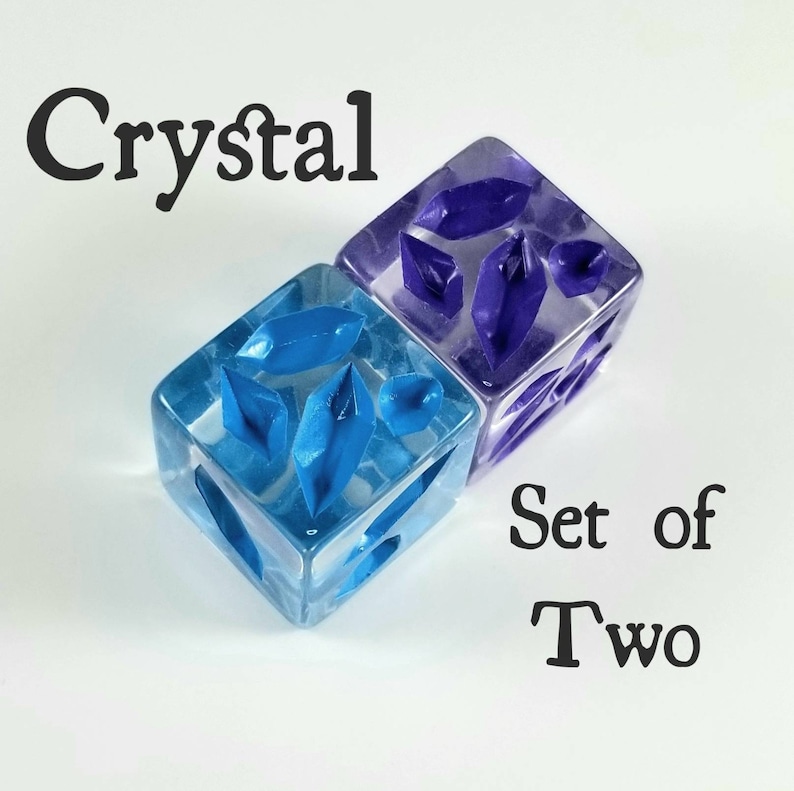 May include: Two dice with crystal-shaped inclusions. One die is blue, the other is purple. The word "Crystal" is above the dice, and "Set of Two" is below. The dice are on a white background.