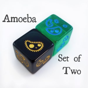 May include: Set of two dice with a black and green color scheme. One die has a gold amoeba design and the other has a blue amoeba design. The dice are labeled "Amoeba Set of Two".