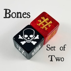 Puede incluir: Conjunto de dos dados, uno negro con una calavera blanca y huesos cruzados, y otro rojo con huesos cruzados dorados. Los dados están etiquetados como "Bones" y "Set of Two".