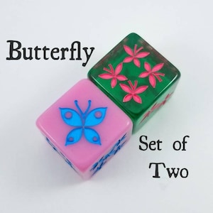 Puede incluir: Conjunto de dos dados con un diseño de mariposa y flores. Los dados son de color rosa y verde con detalles azules y rosas. El texto "Butterfly" y "Set of Two" está impreso en el fondo blanco.