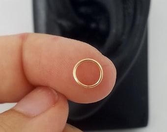May include: A gold hoop earring, shown on a black ear model and held by a hand.