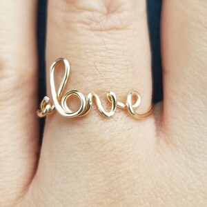 May include: A gold-colored wire ring spelling out the word "love" in cursive script. The ring is delicate and sits on a finger, with a blue background.
