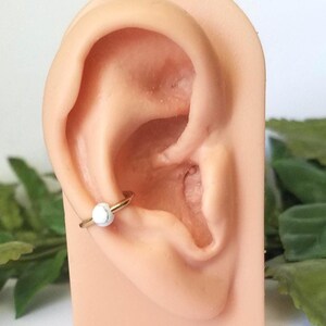 Puede incluir: Un aro de oro para la oreja con una perla de mármol blanco. El aro se lleva en el cartílago superior de la oreja.