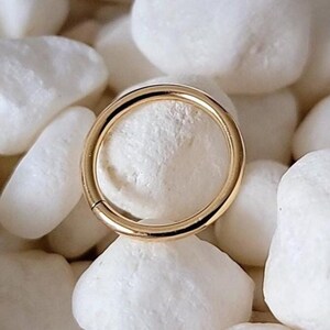 May include: A gold-colored, circular ring is displayed against a backdrop of white, smooth stones. The ring is the focal point, showcasing its polished surface and simple design. The stones provide a contrasting texture and color.