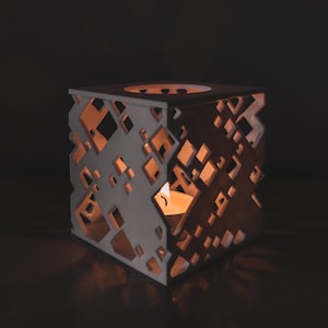 May include: A square, white and brown candle holder with geometric cutouts. The candle holder has a cube shape with diamond-shaped openings that allow the warm light of a lit candle inside to shine through. The candle holder is on a dark surface.