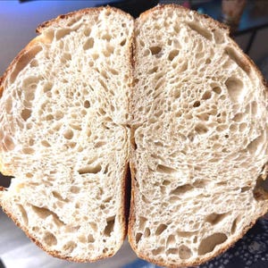 Könnte beinhalten: Nahaufnahme eines in zwei Hälften geschnittenen Brotlaibs, der die innere Textur zeigt. Die Kruste ist goldbraun und das Innere ist hell und luftig mit zahlreichen Löchern. Das Brot scheint frisch gebacken zu sein.