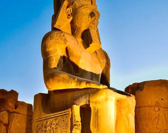 May include: A large, seated statue of a pharaoh with a headdress and a ceremonial beard. The statue is carved from stone and is illuminated by warm light against a blue sky. Intricate carvings adorn the base.